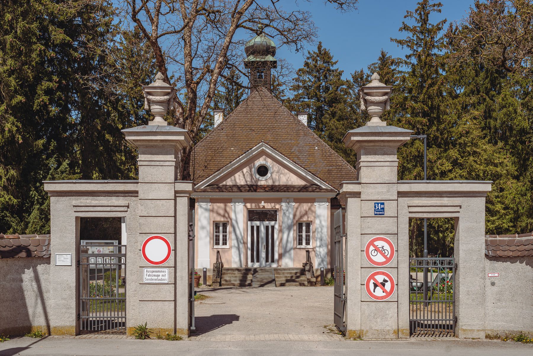 Symmetrical gateway with pale stone pillars and a small building beyond at Obermenzing Cemetery